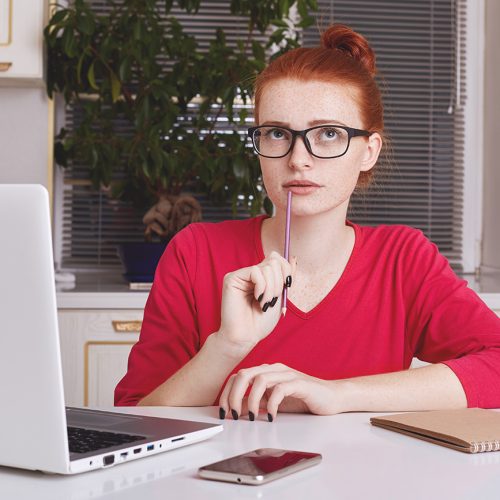 Thoughtful successful young female copywriter tries to get good ideas for new project, puts down notes in diary, works remotely on laptop computer, wants to creat something unsual and creative
