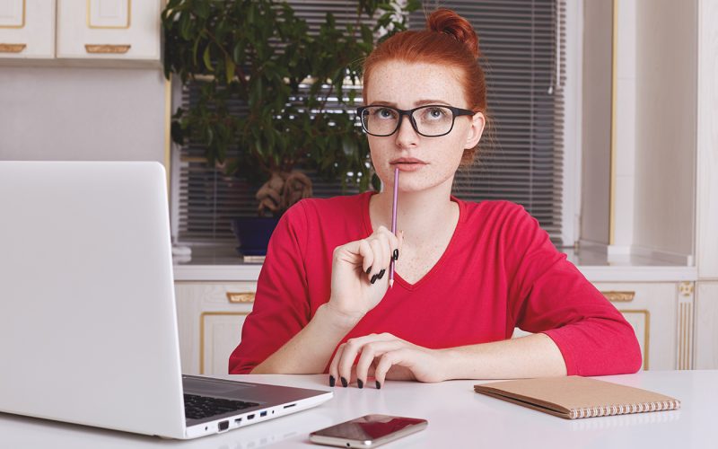 Thoughtful successful young female copywriter tries to get good ideas for new project, puts down notes in diary, works remotely on laptop computer, wants to creat something unsual and creative